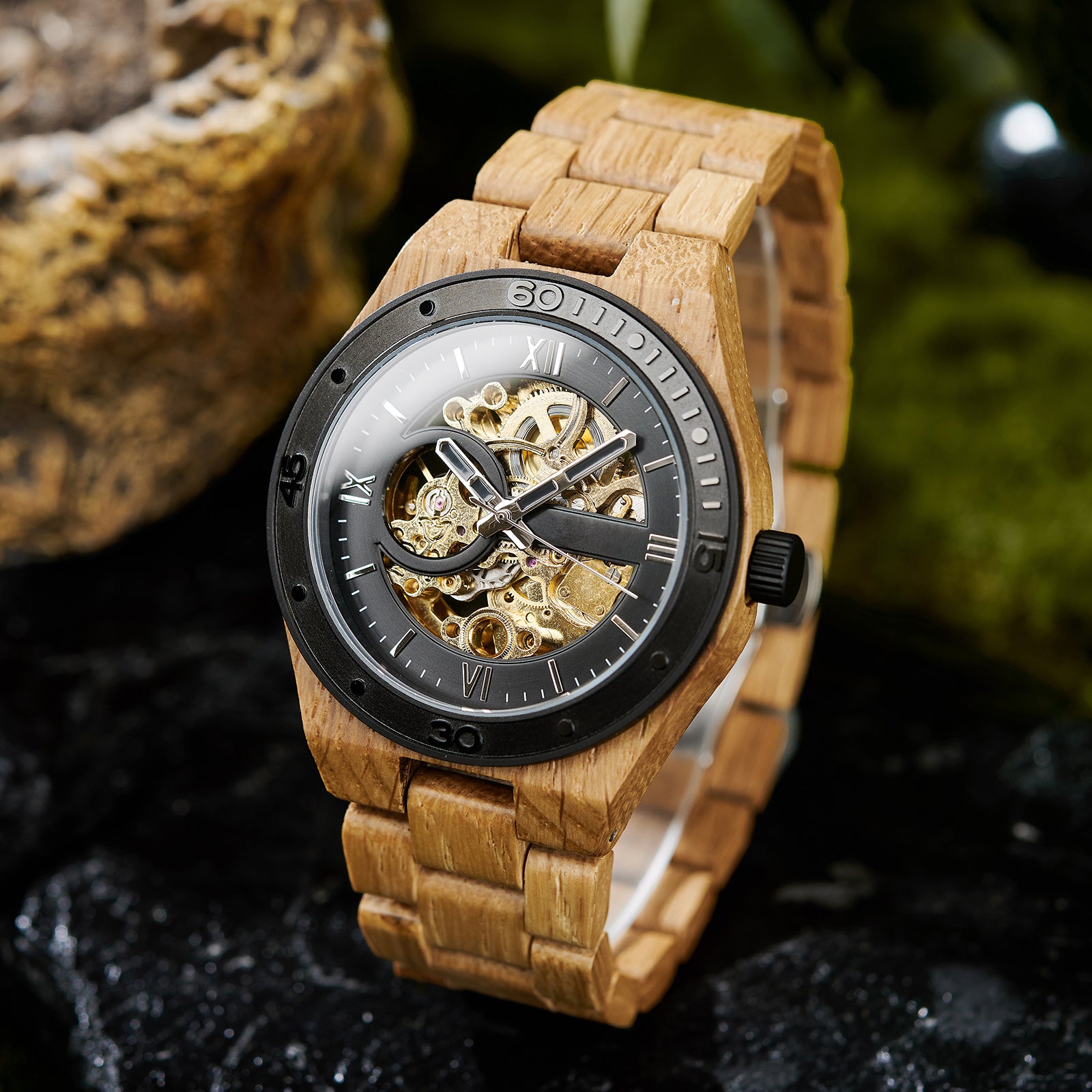 A men's automatic mechanical watch made of oak wood and black stainless steel, featuring a skeleton dial with visible golden mechanical gears, placed on a textured black rock with natural green and brown elements in the background.
