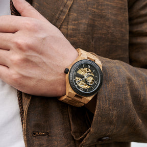 A man wearing a men's automatic mechanical watch made of oak wood and black stainless steel, showcasing a skeleton dial with visible golden mechanical movements, paired with a brown textured jacket, with the watch prominently displayed on the wrist.
