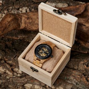 A men's automatic mechanical watch made of oak wood and black stainless steel, placed in a light wooden box with a burlap cushion, set against a textured natural wood background with a gnarled branch.