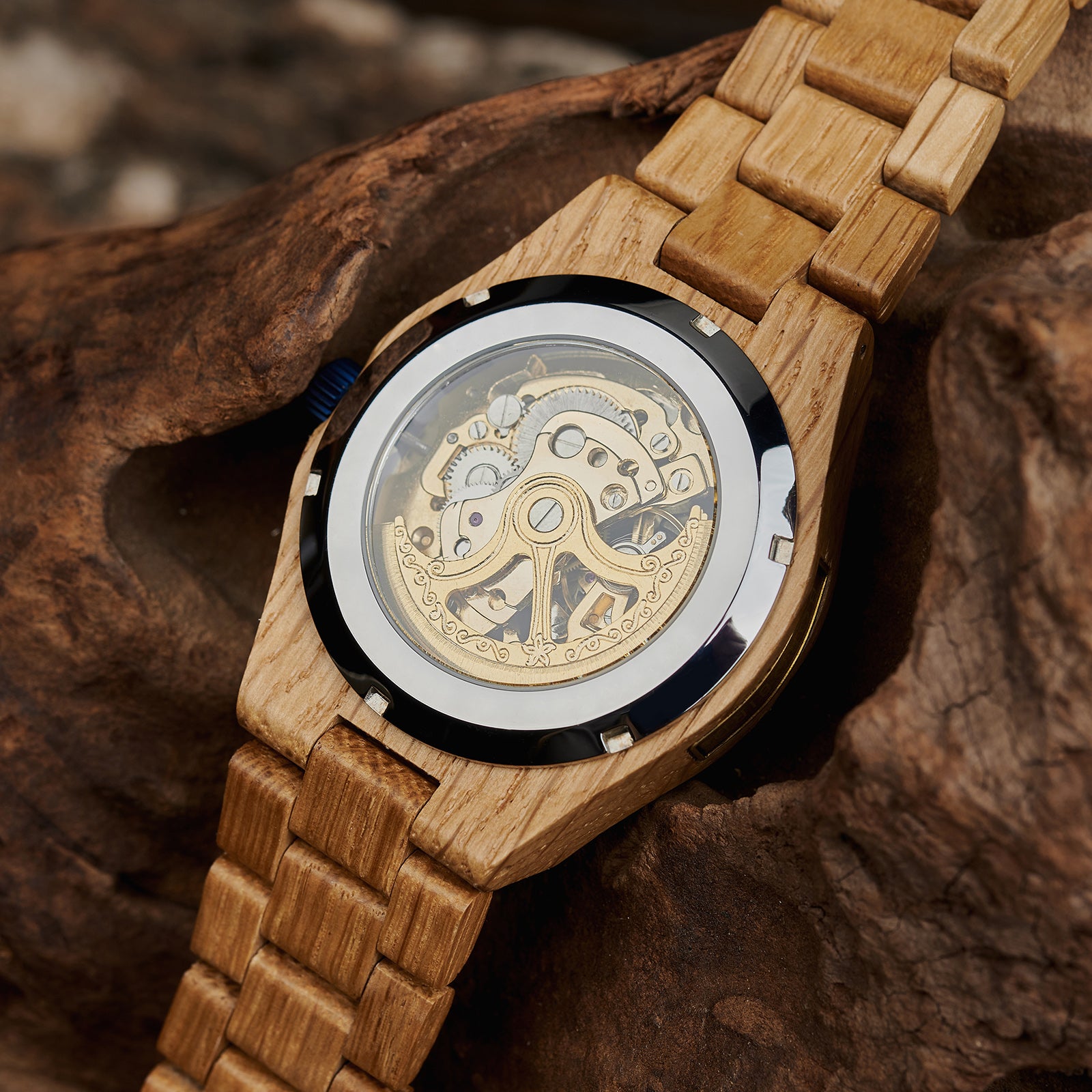 The case back of a men's automatic mechanical watch made of oak wood and blue stainless steel, revealing intricate golden mechanical movements with detailed engravings, placed on textured brown wood against a natural background.