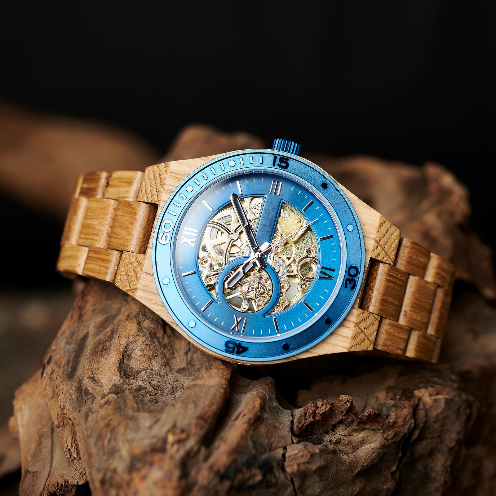A men's automatic mechanical watch made of oak wood and blue stainless steel, featuring a skeleton dial with visible golden mechanical gears, placed on a textured brown rock against a dark background.