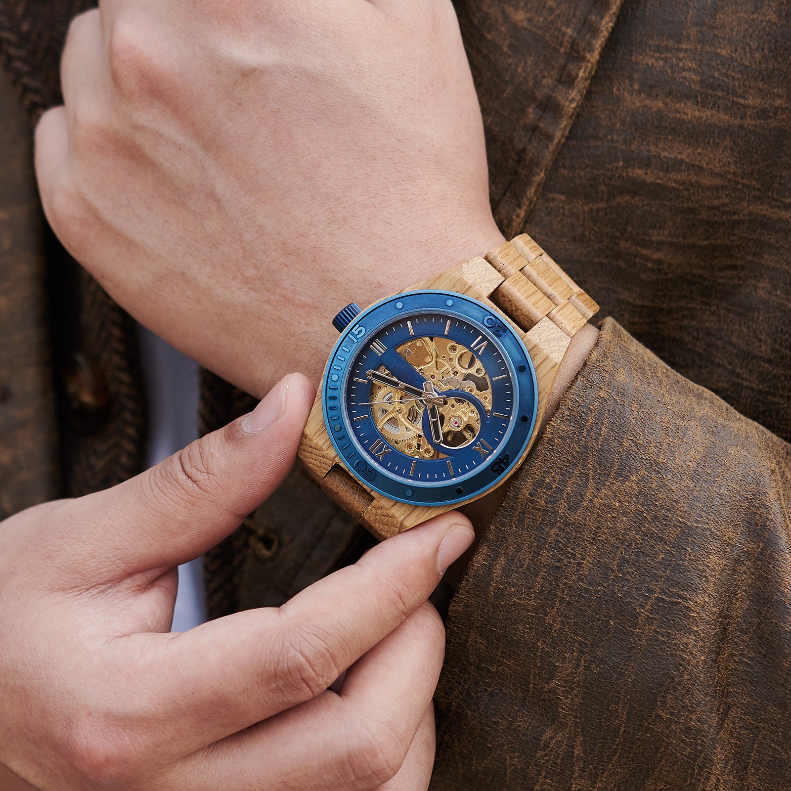 A man wearing a men's automatic mechanical watch made of oak wood and blue stainless steel, showcasing a skeleton dial with visible golden mechanical movements, paired with a brown textured jacket, with one hand adjusting the watch on the wrist.