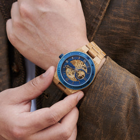 A man wearing a men's automatic mechanical watch made of oak wood and blue stainless steel, showcasing a skeleton dial with visible golden mechanical movements, paired with a brown textured jacket, with one hand adjusting the watch on the wrist.