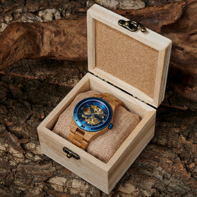 A men's automatic mechanical watch made of oak wood and blue stainless steel, placed in a light wooden box with a cork interior, set against a natural background with rocks, moss, and green plants.