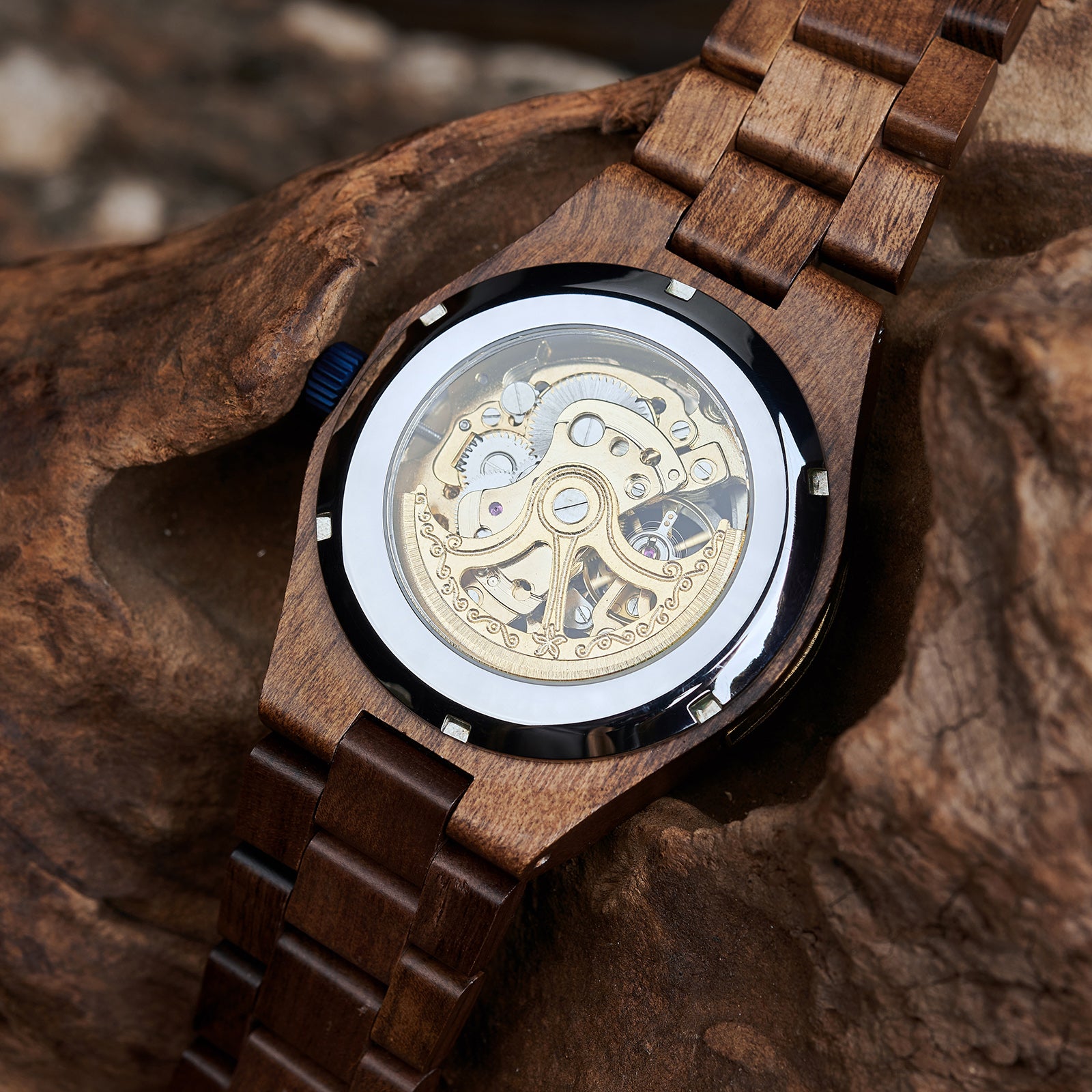 A men's automatic mechanical watch made of walnut wood and black stainless steel, showcasing the intricate engraved movement through a transparent case back, placed on textured wood.
