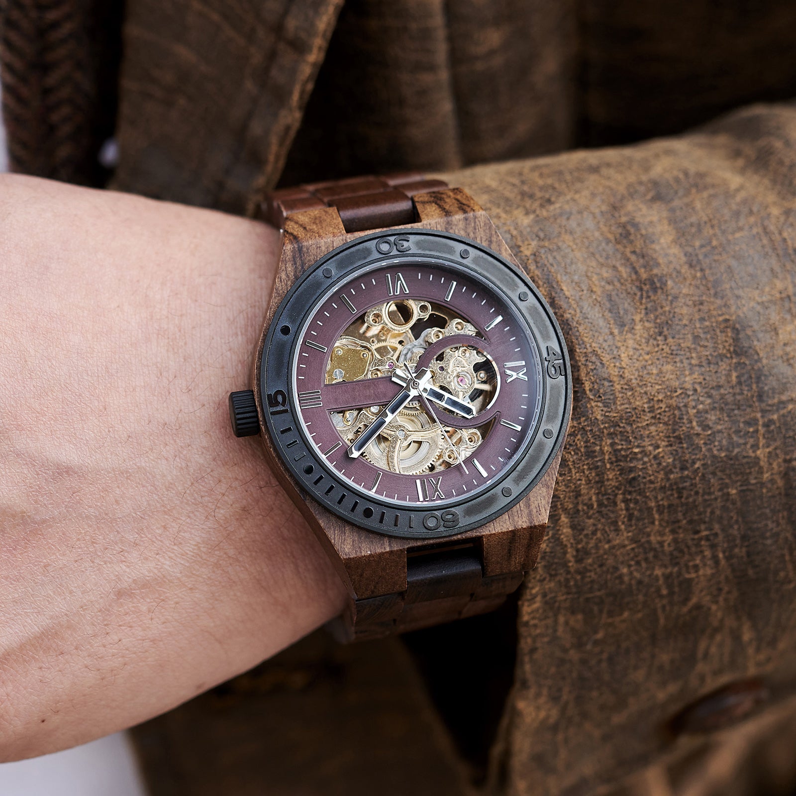 A men's automatic mechanical watch made of walnut wood and black stainless steel, featuring a skeleton dial with intricate movement, worn on a wrist paired with a textured brown jacket.

