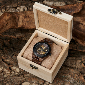 A men's automatic mechanical watch made of walnut wood and black stainless steel, presented in a wooden box with a burlap cushion, placed on textured tree bark with natural wood elements in the background.