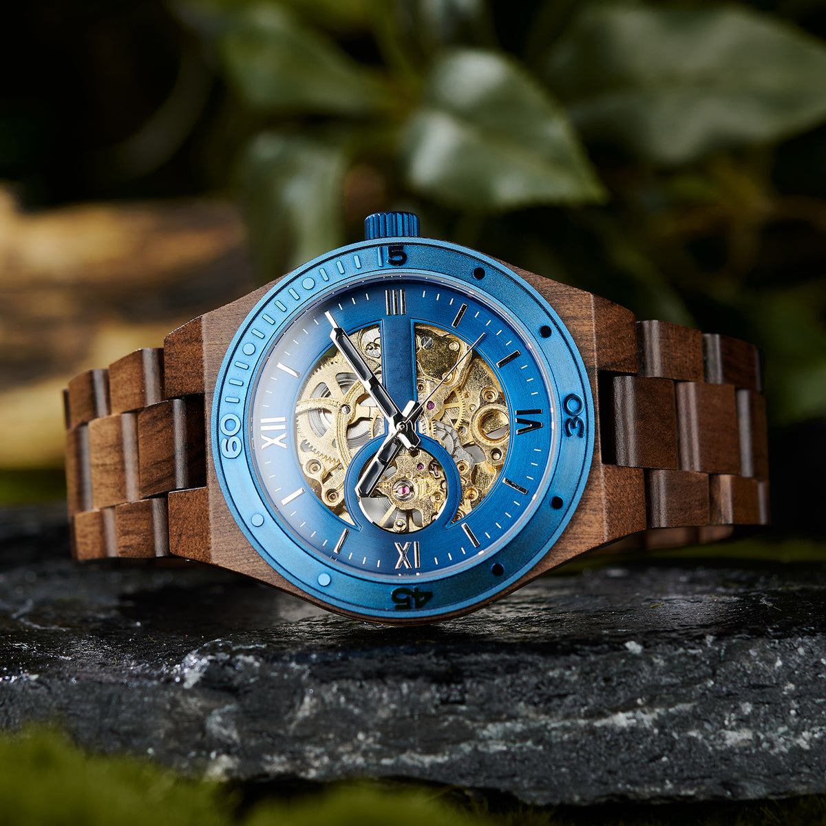 A men's automatic mechanical watch featuring a combination of walnut wood and blue stainless steel, showcasing a skeleton dial with intricate movement, placed on a rock with a natural background.