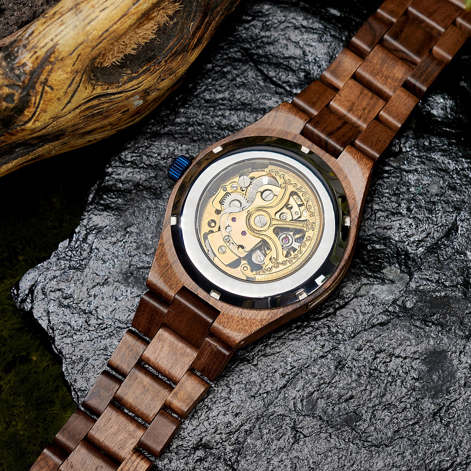 A men's automatic mechanical watch made of walnut wood and blue stainless steel, showcasing the intricate movement through a transparent case back, placed on a textured rock with natural elements in the background.