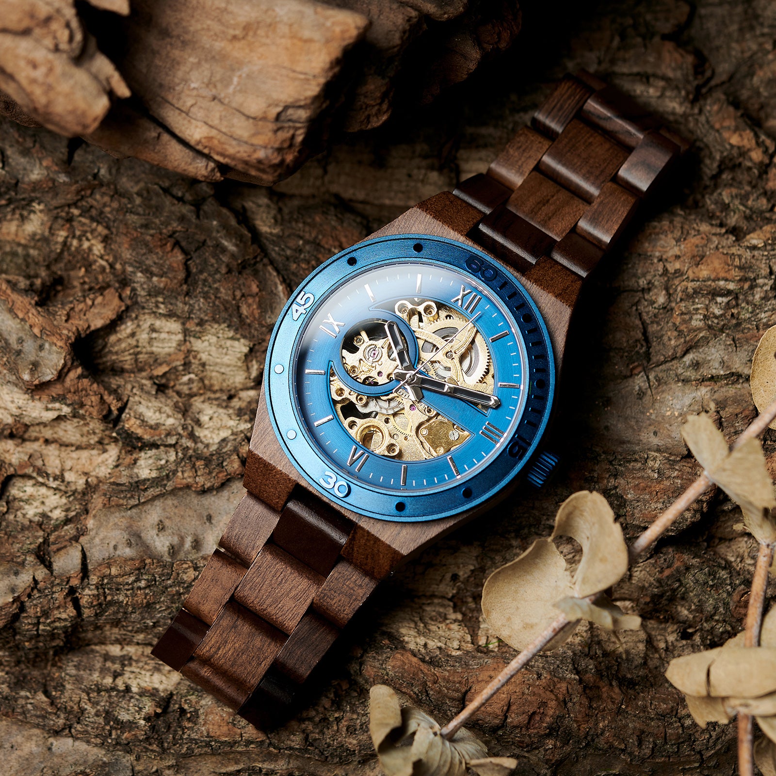A men's automatic mechanical watch made of walnut wood and blue stainless steel, featuring a skeleton dial with intricate movement, placed on a textured tree bark background with natural elements.