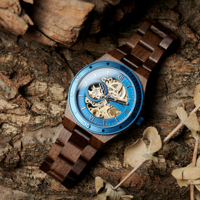 A men's automatic mechanical watch made of walnut wood and blue stainless steel, featuring a skeleton dial with intricate movement, placed on a textured tree bark background with natural elements.