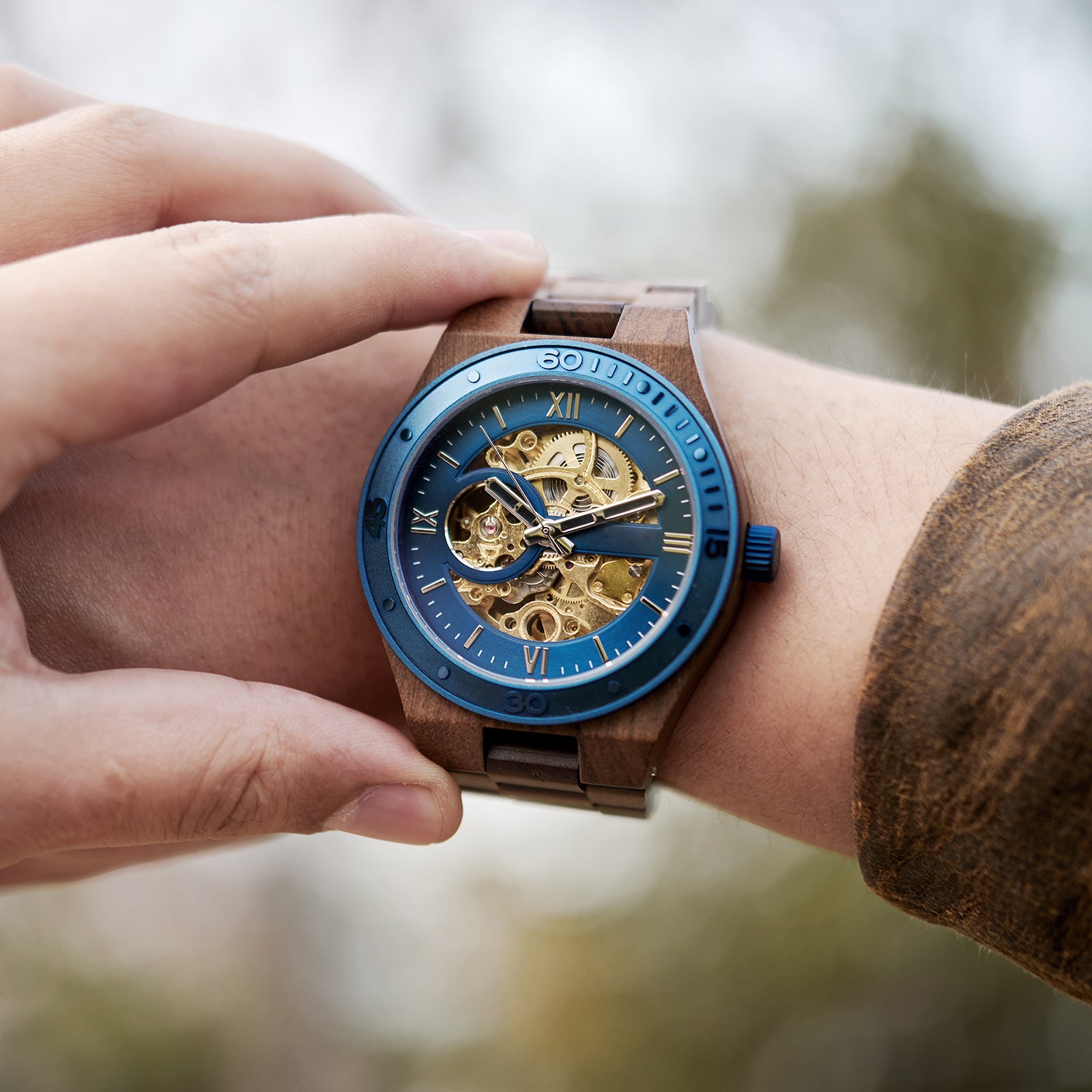 A men's automatic mechanical watch made of walnut wood and blue stainless steel, featuring a skeleton dial with intricate movement, worn on a wrist with a natural outdoor background.

