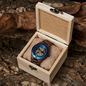A men's automatic mechanical watch made of walnut wood and blue stainless steel, presented in a wooden box with a burlap cushion, placed on a textured tree bark background.
