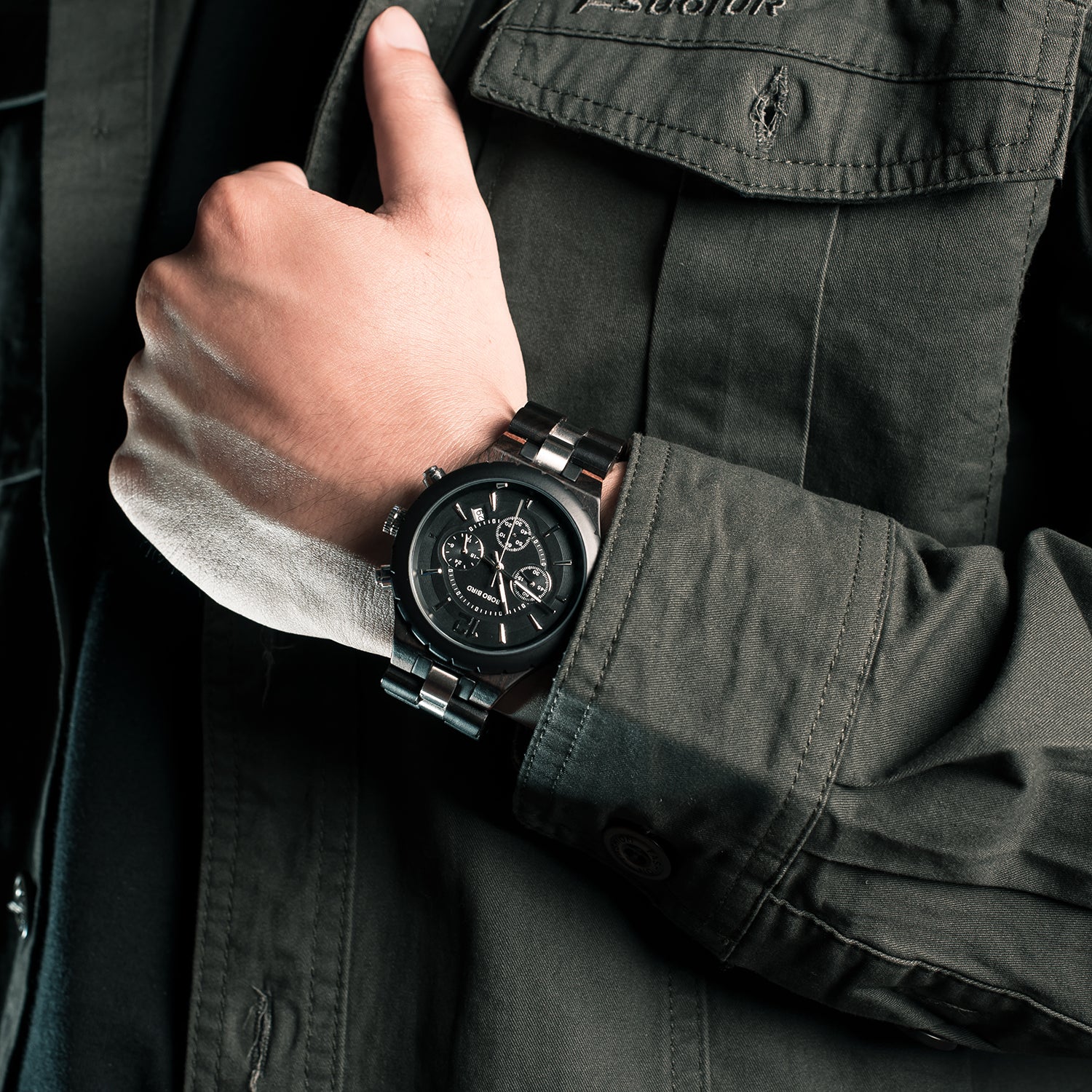 A man wearing a black chronograph watch with a metallic band on his wrist, paired with a dark green jacket, showcasing the watch's design and style.