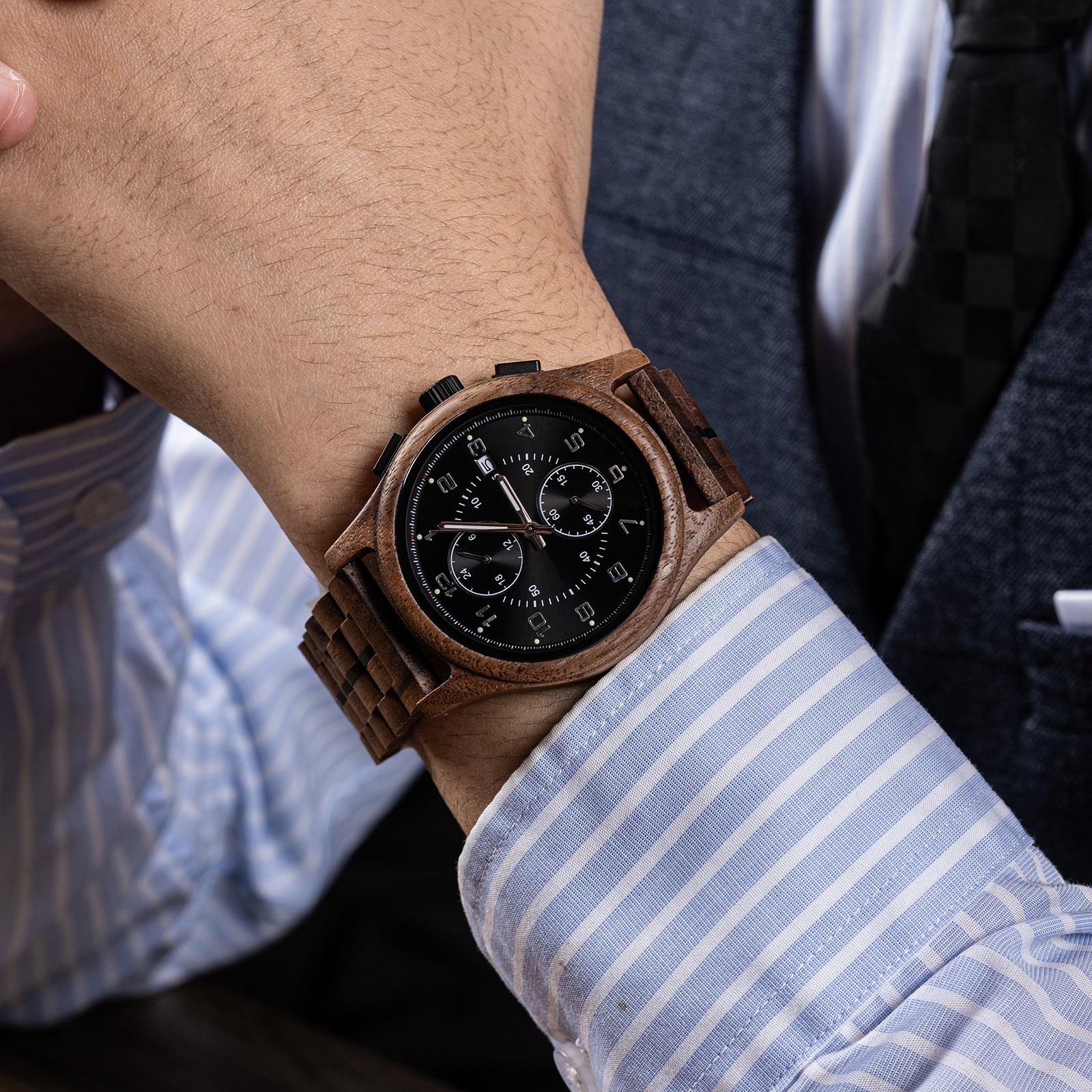 A men's chronograph watch with a brown wooden case and band, featuring a black dial, worn on the wrist of a person dressed in a light blue striped dress shirt and dark suit.