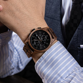 A men's chronograph watch with a brown wooden case and band, featuring a black dial, worn on the wrist of a person dressed in a light blue striped dress shirt and dark suit.