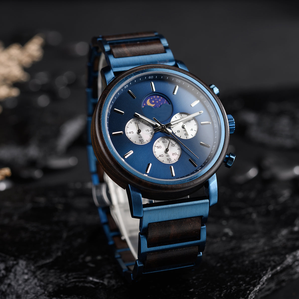 A men's chronograph watch featuring a moon phase complication, blue dial, and a combination of blue stainless steel and wood bracelet, placed on a dark, textured surface.
