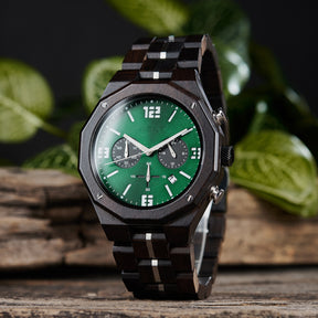 A men's chronograph watch featuring a combination of dark wood and silver-accented bands, a green dial with white numerals and markers, multiple sub-dials, date display, and metallic pushers, set against a background of green foliage and rustic wood.