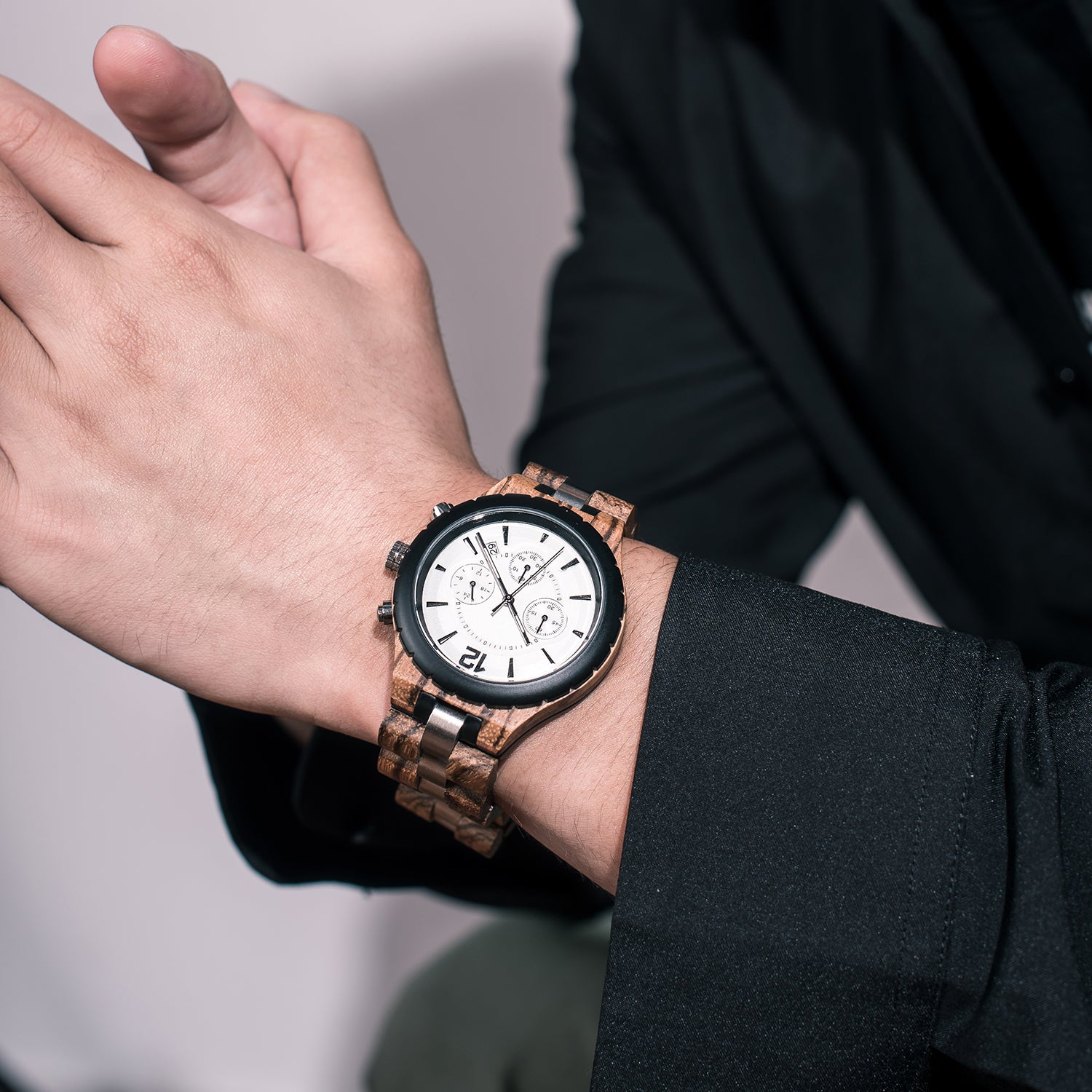 A man wearing a chronograph watch with a combination of light wood and stainless steel bands, featuring a white dial with multiple sub-dials and metallic hour markers, paired with a black jacket.