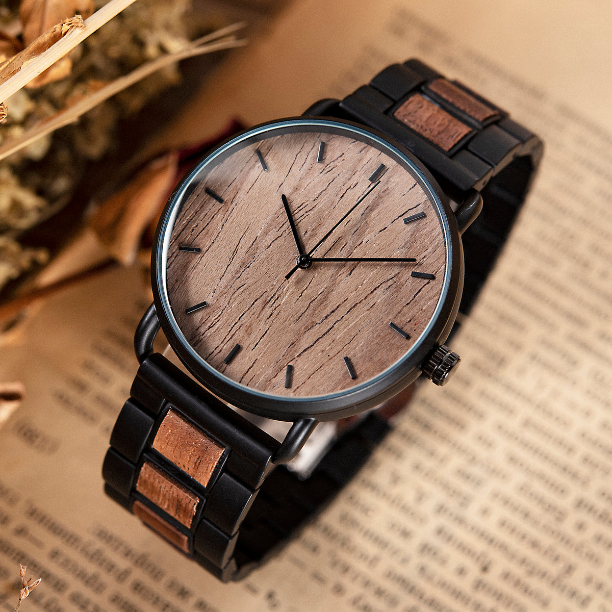 A minimalist men's watch featuring a dark natural wooden dial, black metal and dark wood accent band, presented on a vintage book background with dried foliage to highlight its sleek and natural design aesthetic.