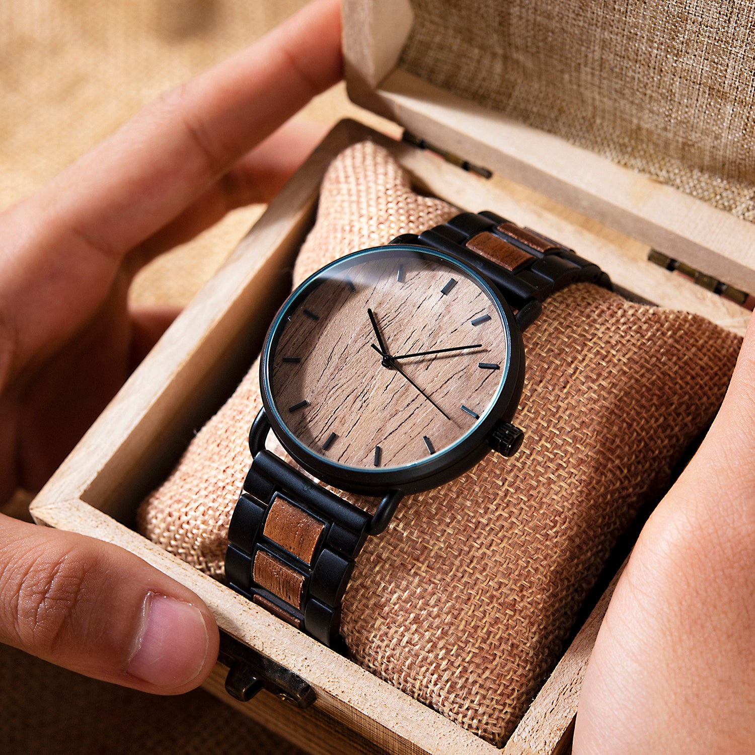 A minimalist men's watch featuring a natural wooden dial, black metal and wood accent band, presented in a wooden box with a burlap pillow to highlight its sleek and natural design aesthetic.