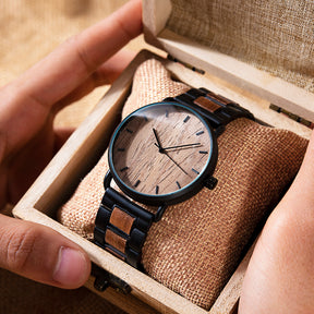A minimalist men's watch featuring a natural wooden dial, black metal and wood accent band, presented in a wooden box with a burlap pillow to highlight its sleek and natural design aesthetic.