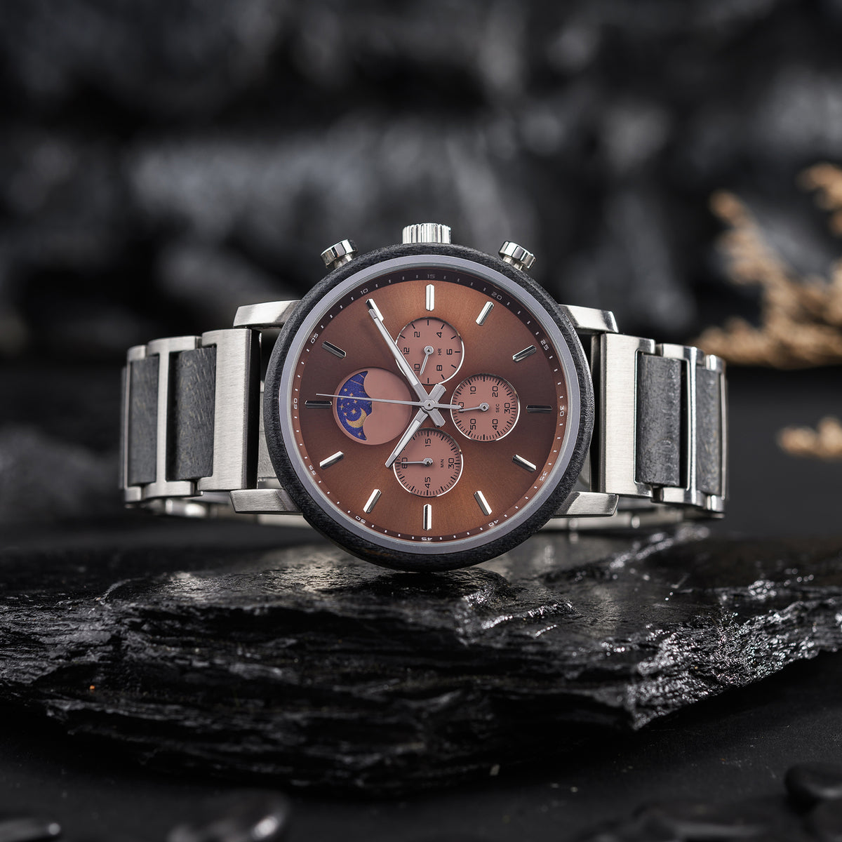 A men's multi-function watch featuring a moon phase complication and chronograph sub-dials, with a brown dial, stainless steel and gray strap, placed on black slate against a dark, textured background.