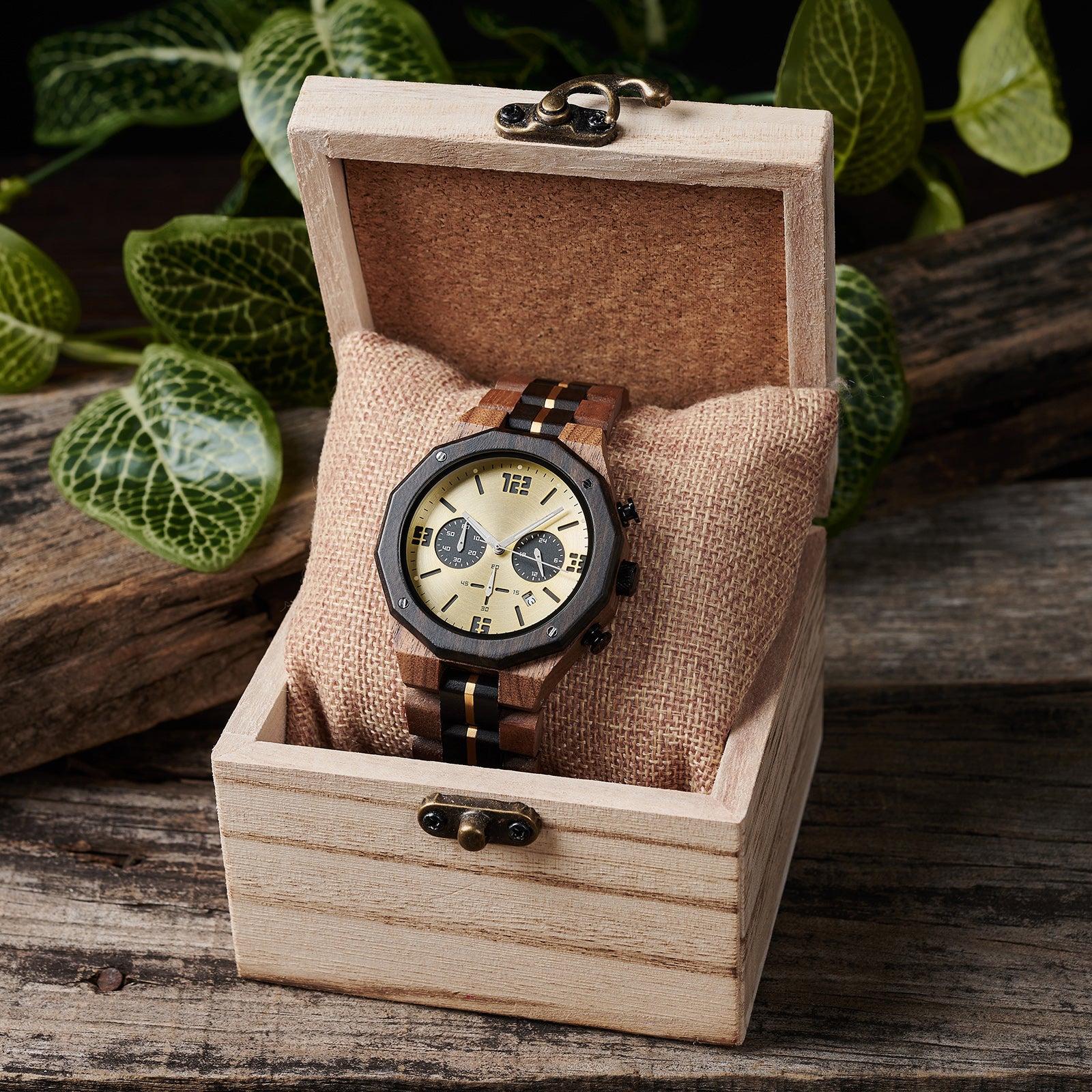 A men's chronograph watch featuring a combination of multicolored wood and gold-accented bands, a gold dial with bold numerals, multiple sub-dials, date display, and metallic pushers, placed on a burlap pillow inside a light wooden box with vintage clasps, set against a background of rustic wood and green foliage.