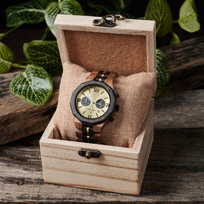 A men's chronograph watch featuring a combination of multicolored wood and gold-accented bands, a gold dial with bold numerals, multiple sub-dials, date display, and metallic pushers, placed on a burlap pillow inside a light wooden box with vintage clasps, set against a background of rustic wood and green foliage.