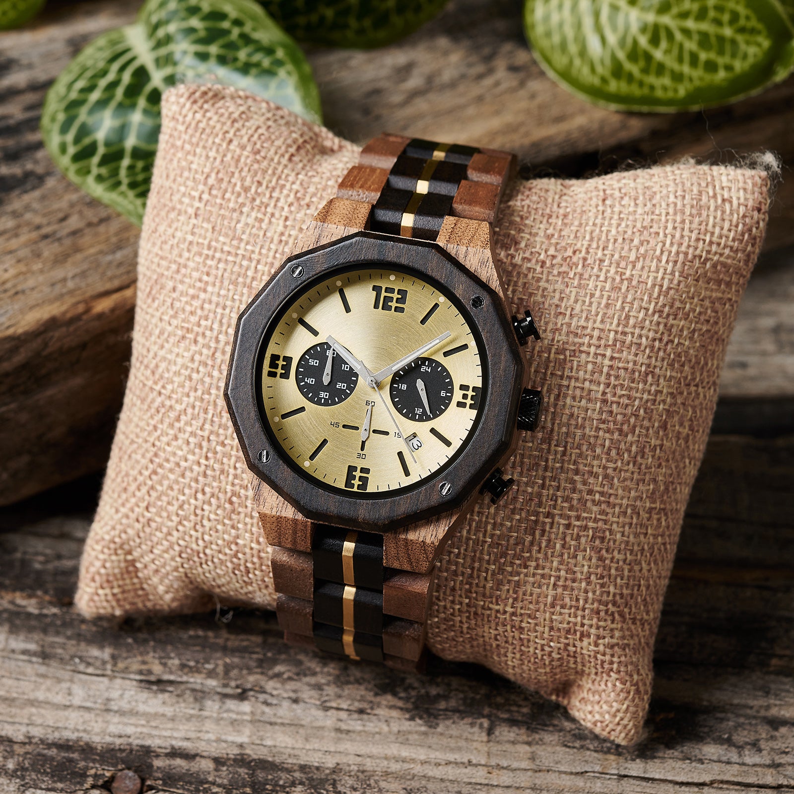 A men's chronograph watch featuring a combination of multicolored wood and gold-accented bands, a gold dial with bold numerals, multiple sub-dials, date display, and metallic pushers, placed on a burlap pillow against a background of rustic wood and green foliage.