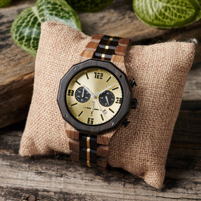 A men's chronograph watch featuring a combination of multicolored wood and gold-accented bands, a gold dial with bold numerals, multiple sub-dials, date display, and metallic pushers, placed on a burlap pillow against a background of rustic wood and green foliage.