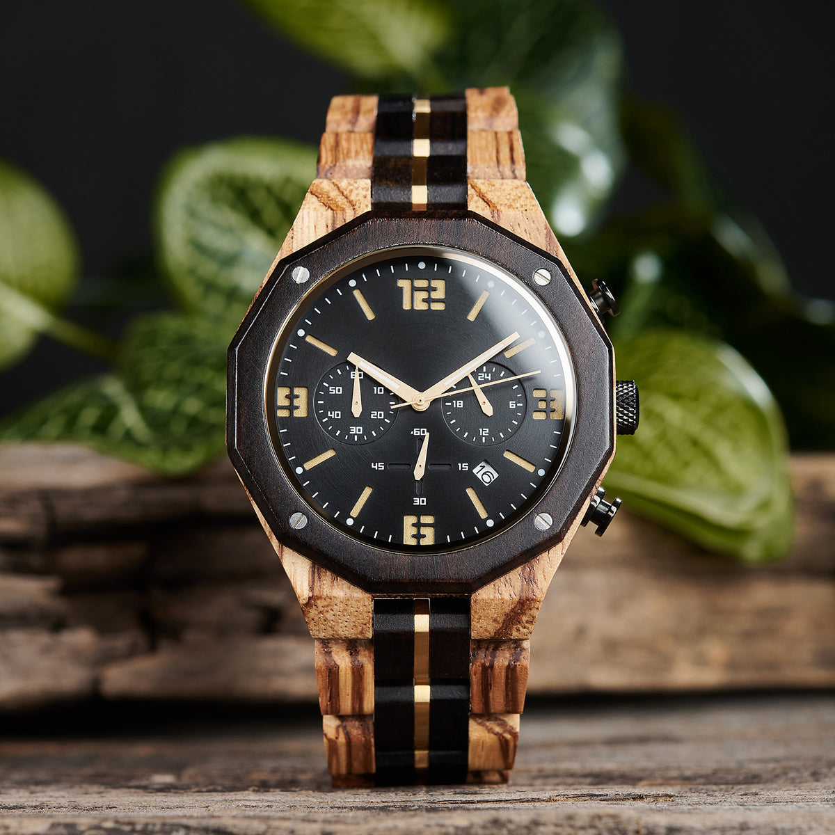 A men's chronograph watch featuring a combination of multicolored wood and gold-accented bands, a black dial with gold numerals and markers, multiple sub-dials, date display, and metallic pushers, set against a background of green foliage and rustic wood.