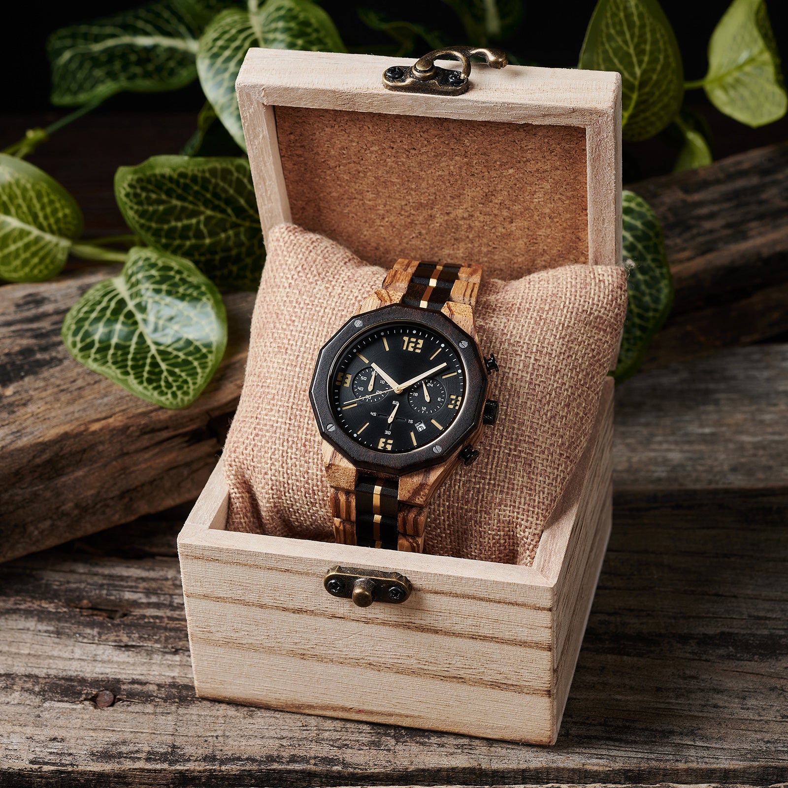 A men's chronograph watch featuring a combination of multicolored wood and gold-accented bands, a black dial with gold numerals and markers, multiple sub-dials, date display, and metallic pushers, placed on a burlap pillow inside a light wooden box with vintage clasps, set against a background of rustic wood and green foliage.