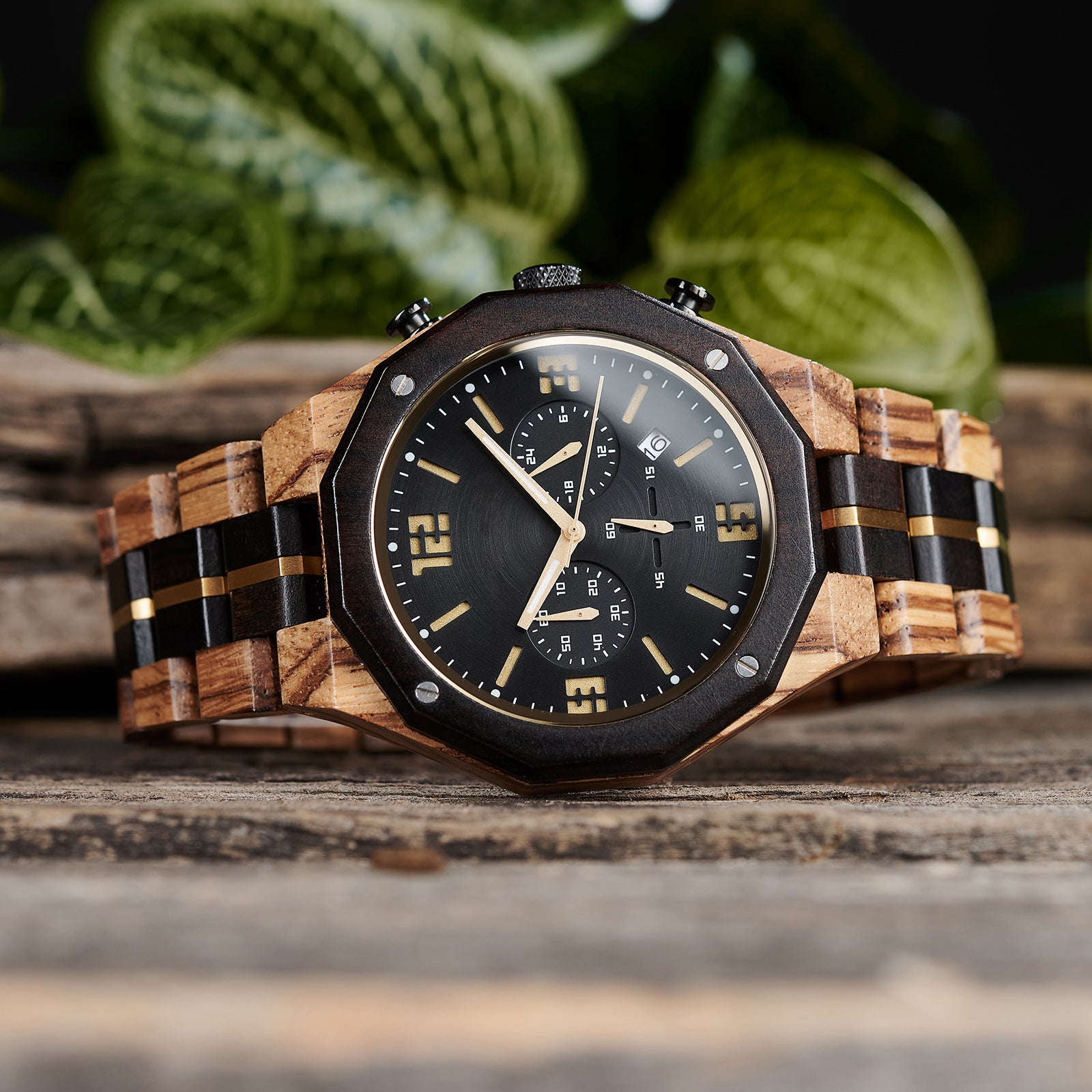 A men's chronograph watch featuring a combination of multicolored wood and gold-accented bands, a black dial with gold numerals and markers, multiple sub-dials, date display, and metallic pushers, shown from a side angle against a background of green foliage and rustic wood.