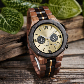 A men's chronograph watch featuring a combination of multicolored wood and gold-accented bands, a gold dial with bold numerals, multiple sub-dials, date display, and metallic pushers, set against a background of green foliage and rustic wood.