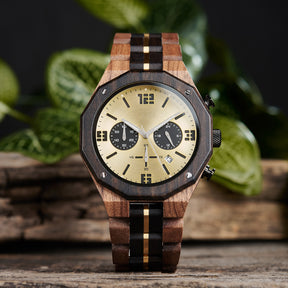 A men's chronograph watch featuring a combination of multicolored wood and gold-accented bands, a gold dial with bold numerals, multiple sub-dials, date display, and metallic pushers, set against a background of green foliage and rustic wood.