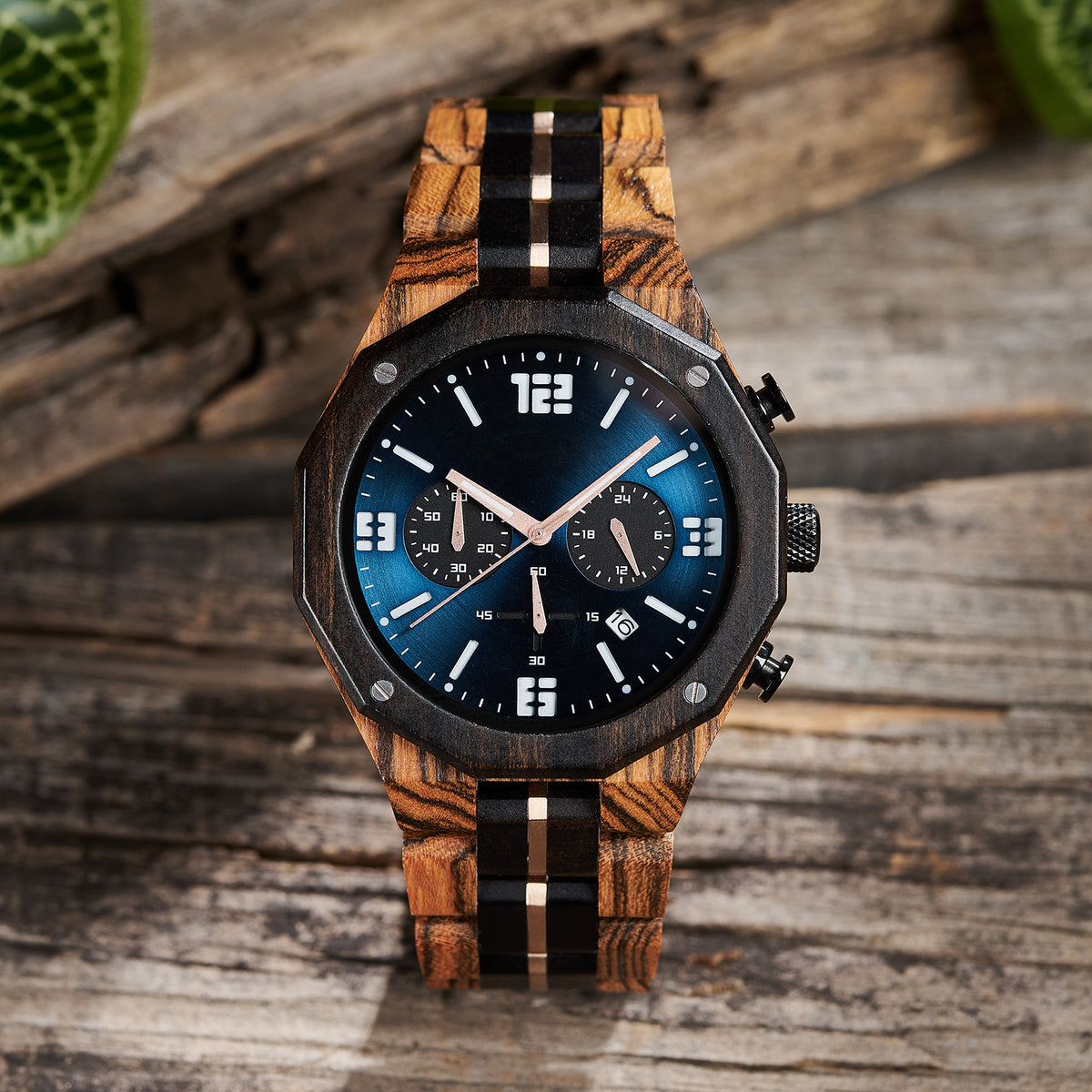 A men's chronograph watch featuring a combination of multicolored wood and rose gold-accented bands, a blue dial with white numerals and markers, multiple sub-dials, date display, and metallic pushers, set against a background of rustic wood and green foliage.