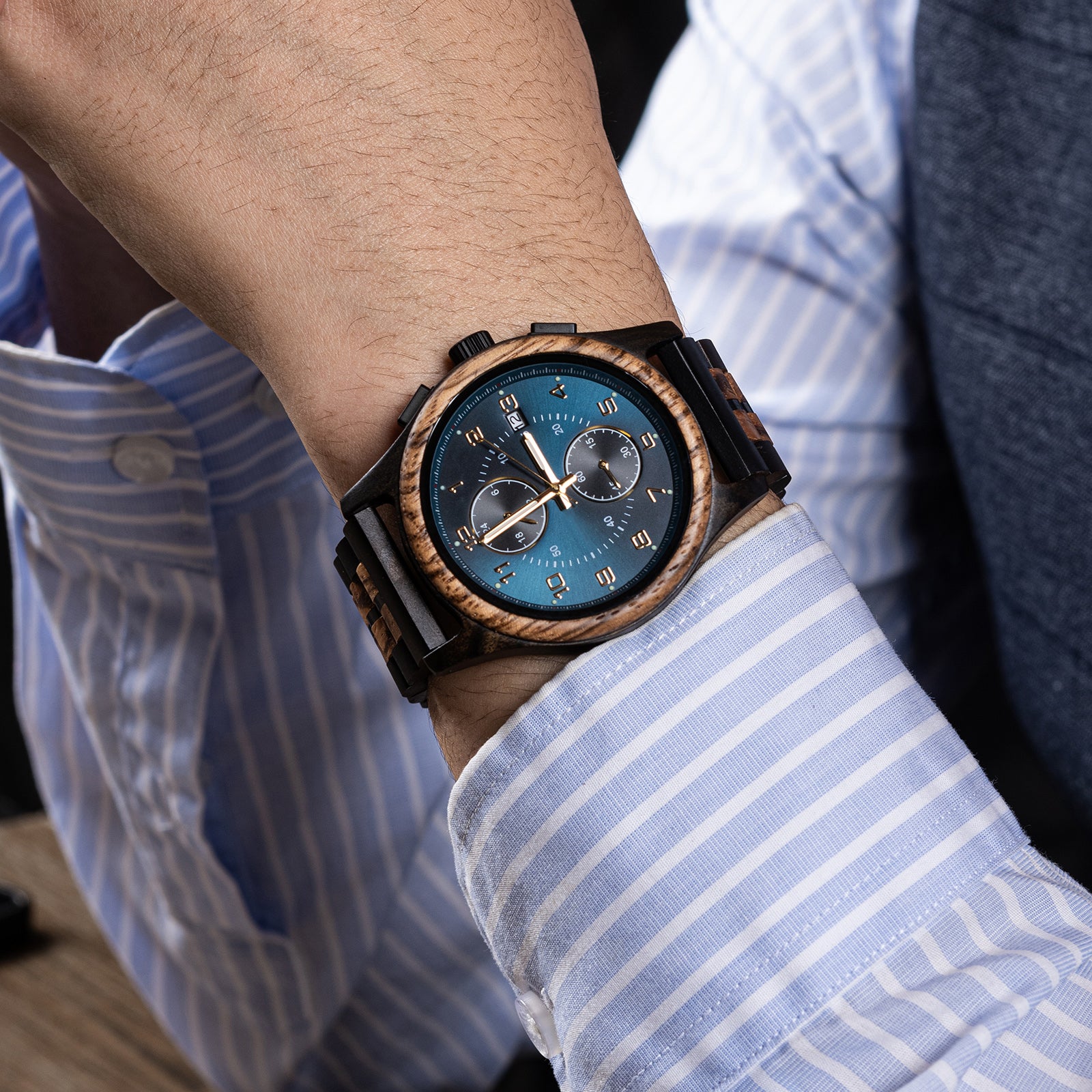 A men's chronograph watch with wooden and black metal details, featuring a teal dial, worn on the wrist of a person in a light blue striped dress shirt.
