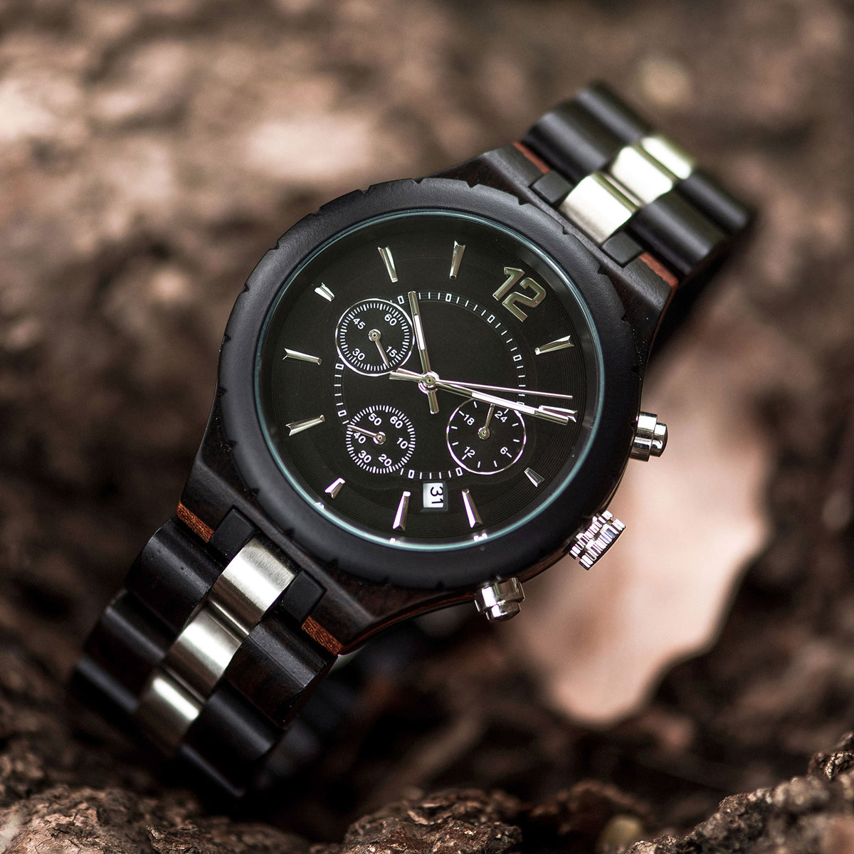 A men's chronograph watch with a combination of dark wood and stainless steel bands, featuring a black dial with multiple sub-dials, date display, and metallic hour markers, placed on a textured bark surface.