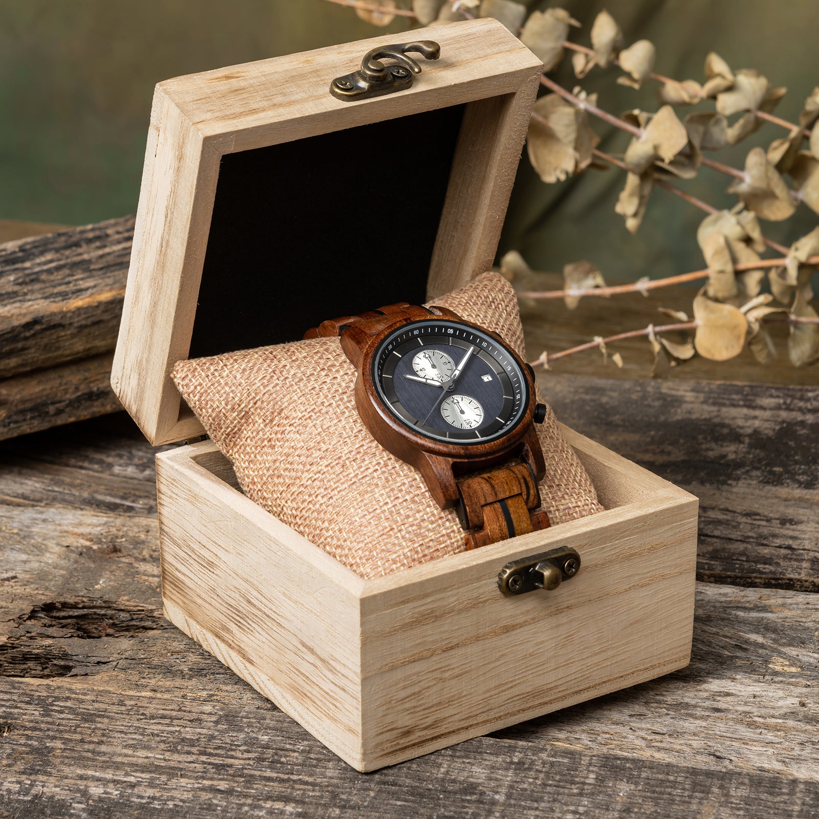 A sophisticated men's wooden chronograph watch featuring a dark gray dial, multiple sub-dials, and a date function, presented in a wooden box with a burlap pillow against a rustic wood background to emphasize its elegant and natural packaging.