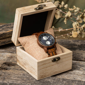 A sophisticated men's wooden chronograph watch featuring a dark gray dial, multiple sub-dials, and a date function, presented in a wooden box with a burlap pillow against a rustic wood background to emphasize its elegant and natural packaging.