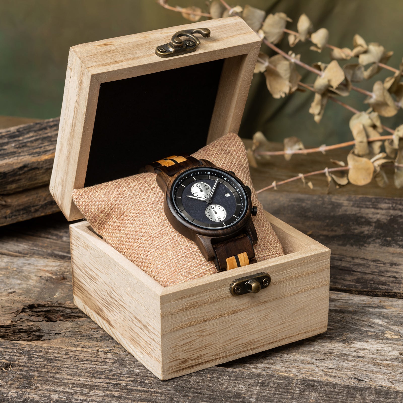 A sophisticated men's wooden chronograph watch featuring a two-tone wood band, dark gray dial, multiple sub-dials, and a date function, presented in a wooden box with a burlap pillow against a rustic wood background to emphasize its elegant and natural packaging.