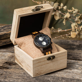 A sophisticated men's wooden chronograph watch featuring a two-tone wood band, dark gray dial, multiple sub-dials, and a date function, presented in a wooden box with a burlap pillow against a rustic wood background to emphasize its elegant and natural packaging.