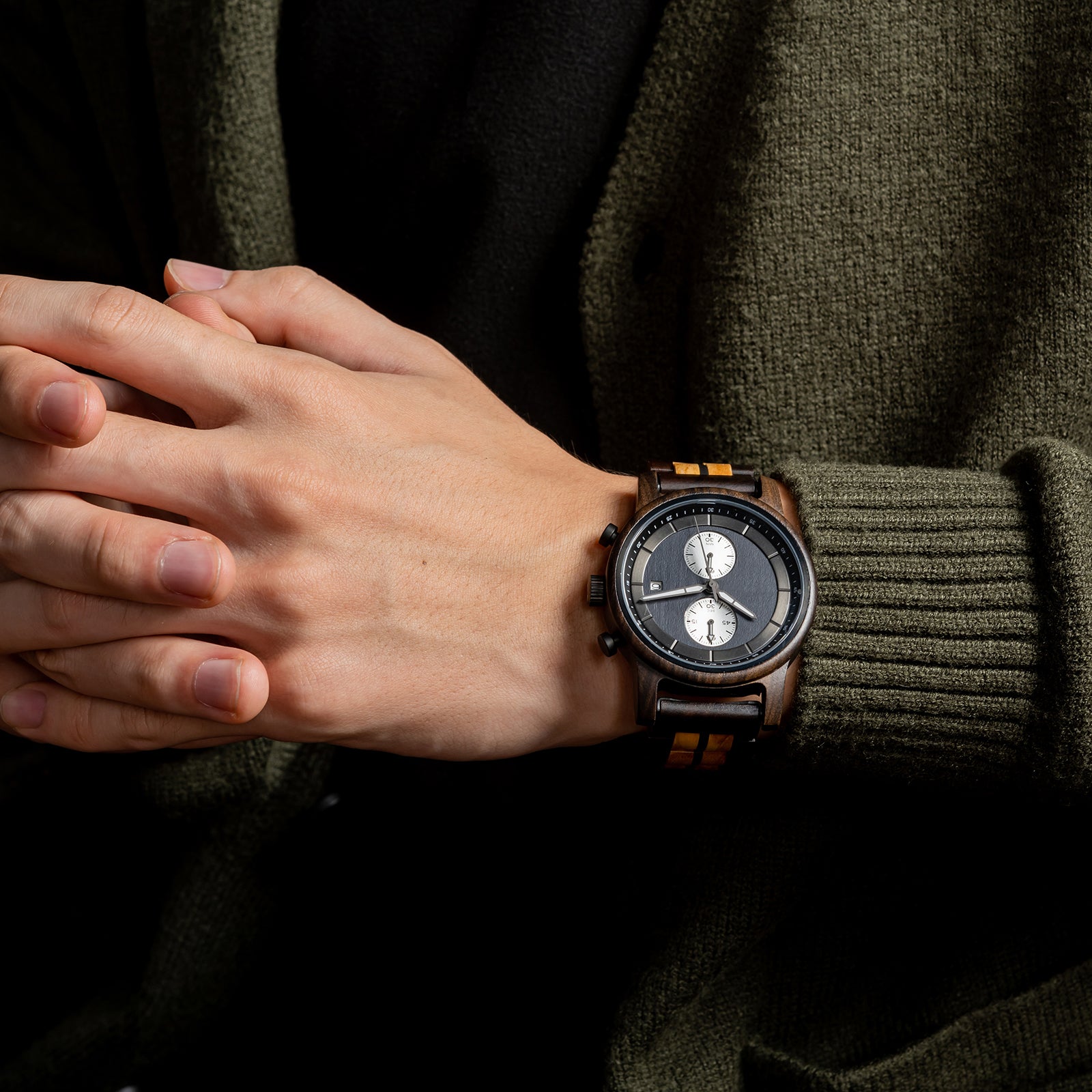 A stylish men's wooden chronograph watch with a two-tone wood band, showcased on a wrist paired with an olive green sweater, highlighting its sophisticated and versatile aesthetic in a lifestyle context.