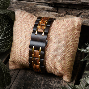 A detailed view of a men's wooden watch band featuring two-tone wood and gold accents, presented on a burlap pillow with a rustic wood and green foliage background to highlight its natural and refined design.