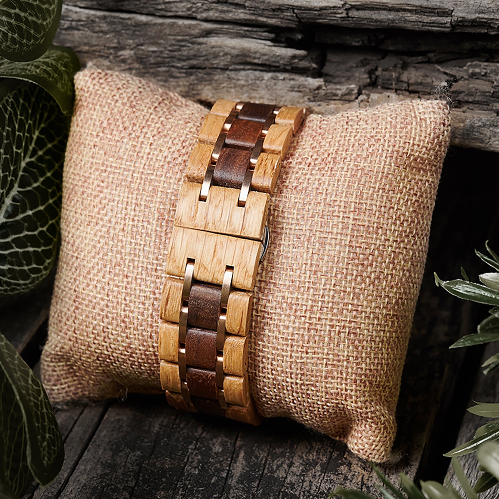 A detailed view of a men's wooden watch band featuring two-tone wood and rose gold accents, presented on a burlap pillow with a rustic wood and green foliage background to highlight its natural and refined design.