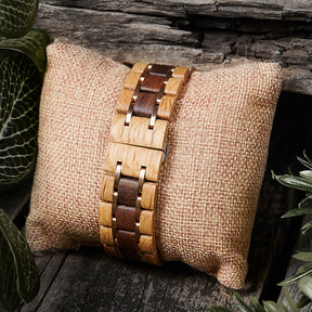 A detailed view of a men's wooden watch band featuring two-tone wood and rose gold accents, presented on a burlap pillow with a rustic wood and green foliage background to highlight its natural and refined design.