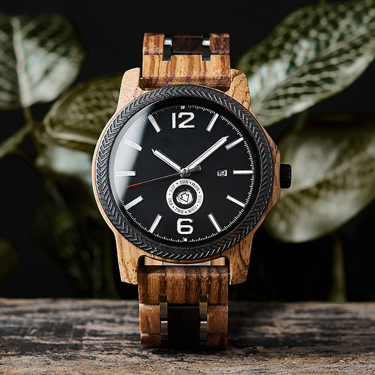 A stylish men's wooden watch featuring a black dial, date and day display, textured bezel, and natural wood grain band, presented against a backdrop of green foliage to highlight its rugged yet sophisticated aesthetic