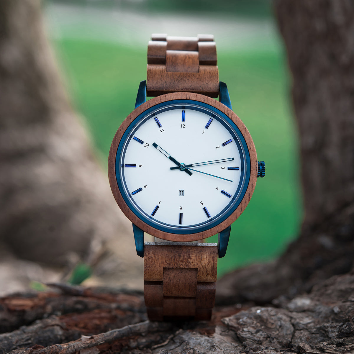 A stylish men's watch featuring a white dial with blue accents, light brown wooden band, and blue metal details, presented on tree bark amidst a green outdoor background to highlight its natural and modern design aesthetic.