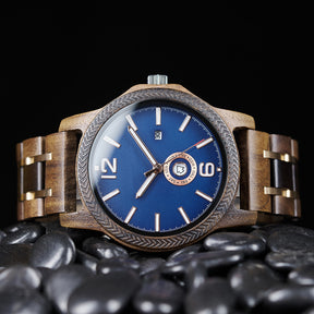 A sophisticated men's wooden watch featuring a blue dial, date and day display, textured bezel, and rose gold-accented band, presented on black pebbles against a dark background to highlight its elegant and refined aesthetic.